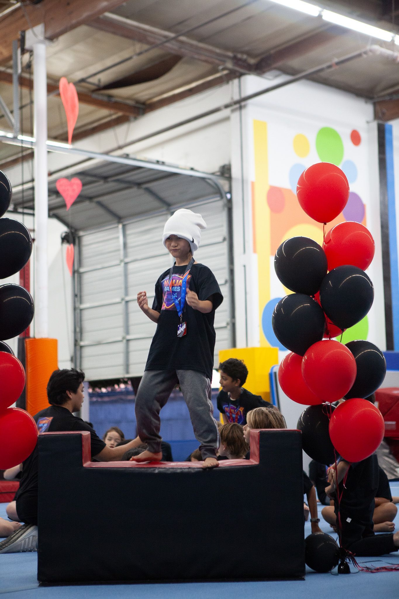 A child in a chef hat stands on a black platform, surrounded by balloons; children in matching shirts watch.