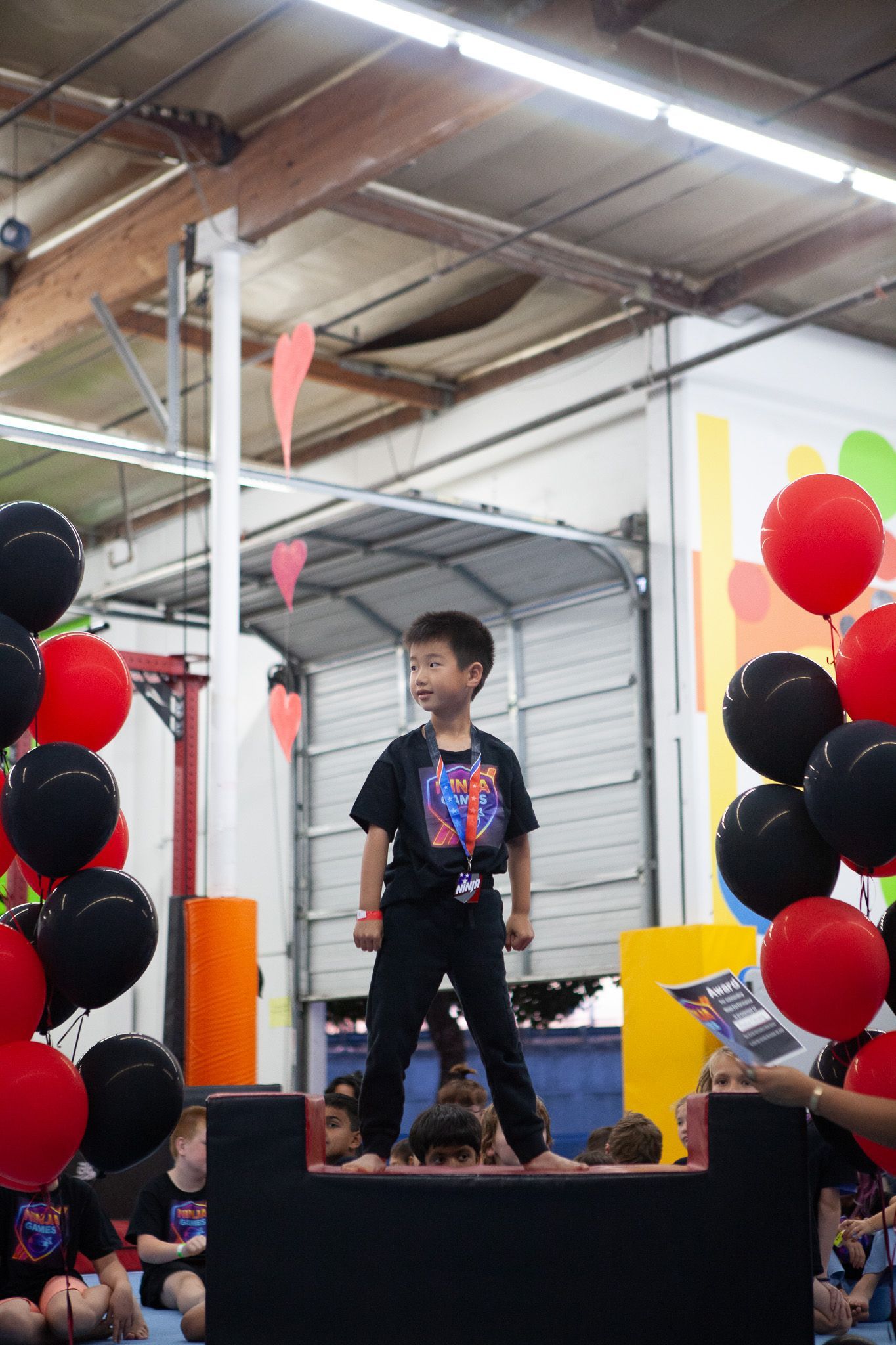 Boy standing on a platform, smiling. Red and black balloons flank him in a gym.