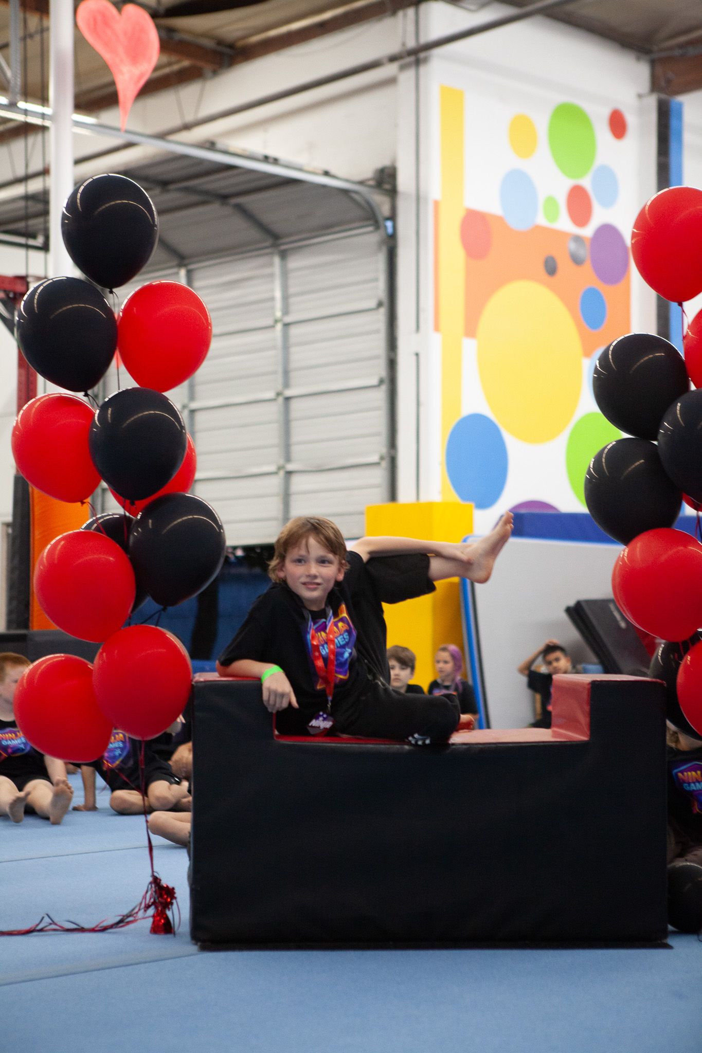 Child balancing on black block, smiling, surrounded by red and black balloons, inside a gym.