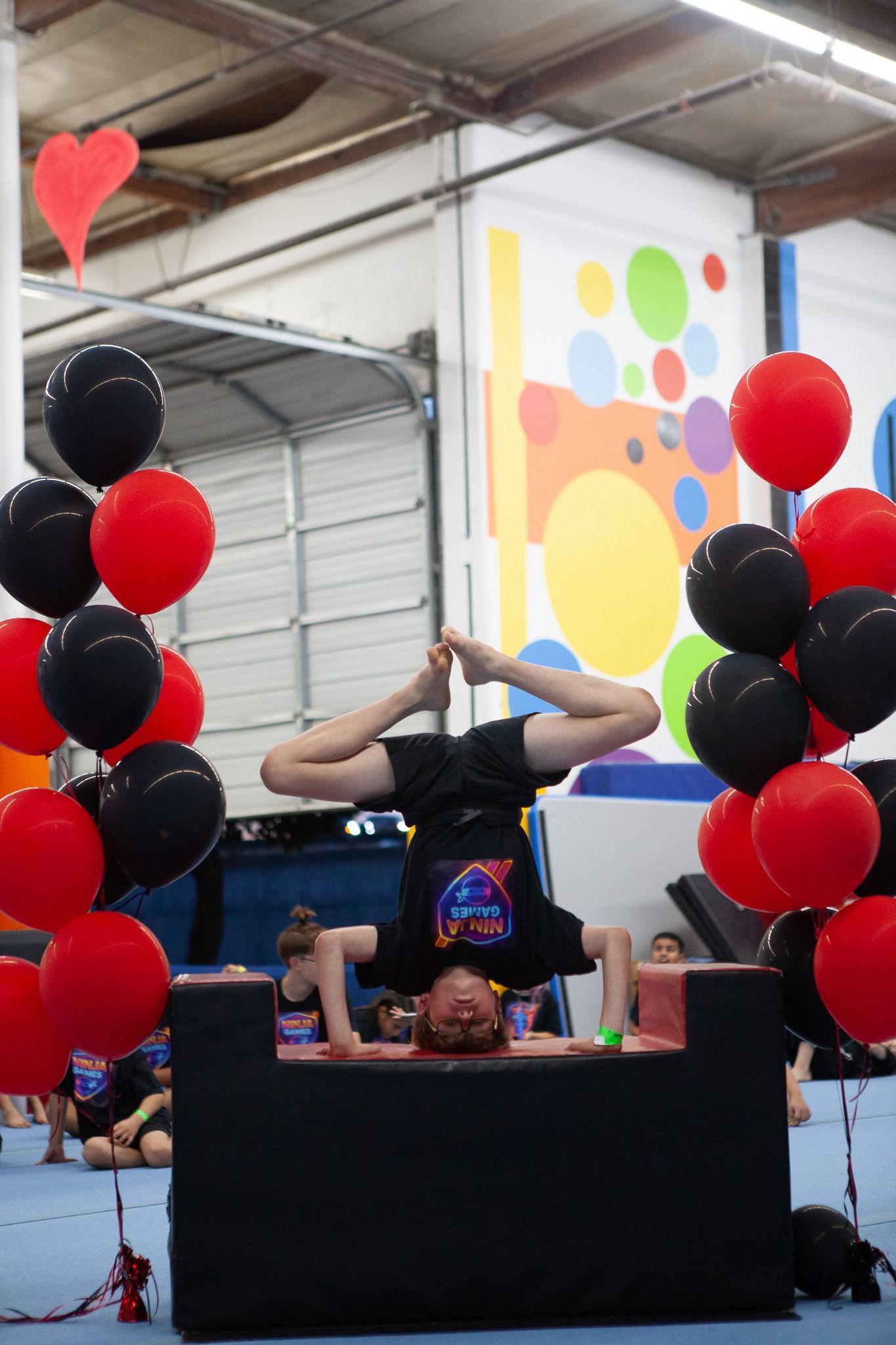 Boy doing a headstand on a black platform, surrounded by red and black balloons, in a gym.