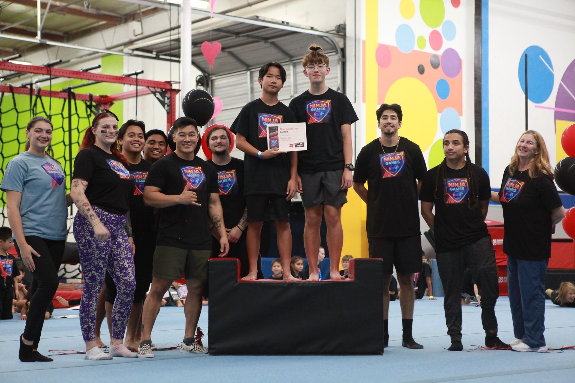 Group stands on podium, holding certificate in an indoor gym. Colorful mural and balloons in background.