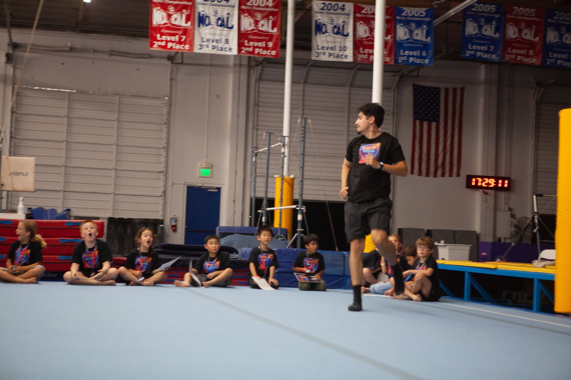 Gymnast running in front of a group of children seated on a blue mat inside a gym.