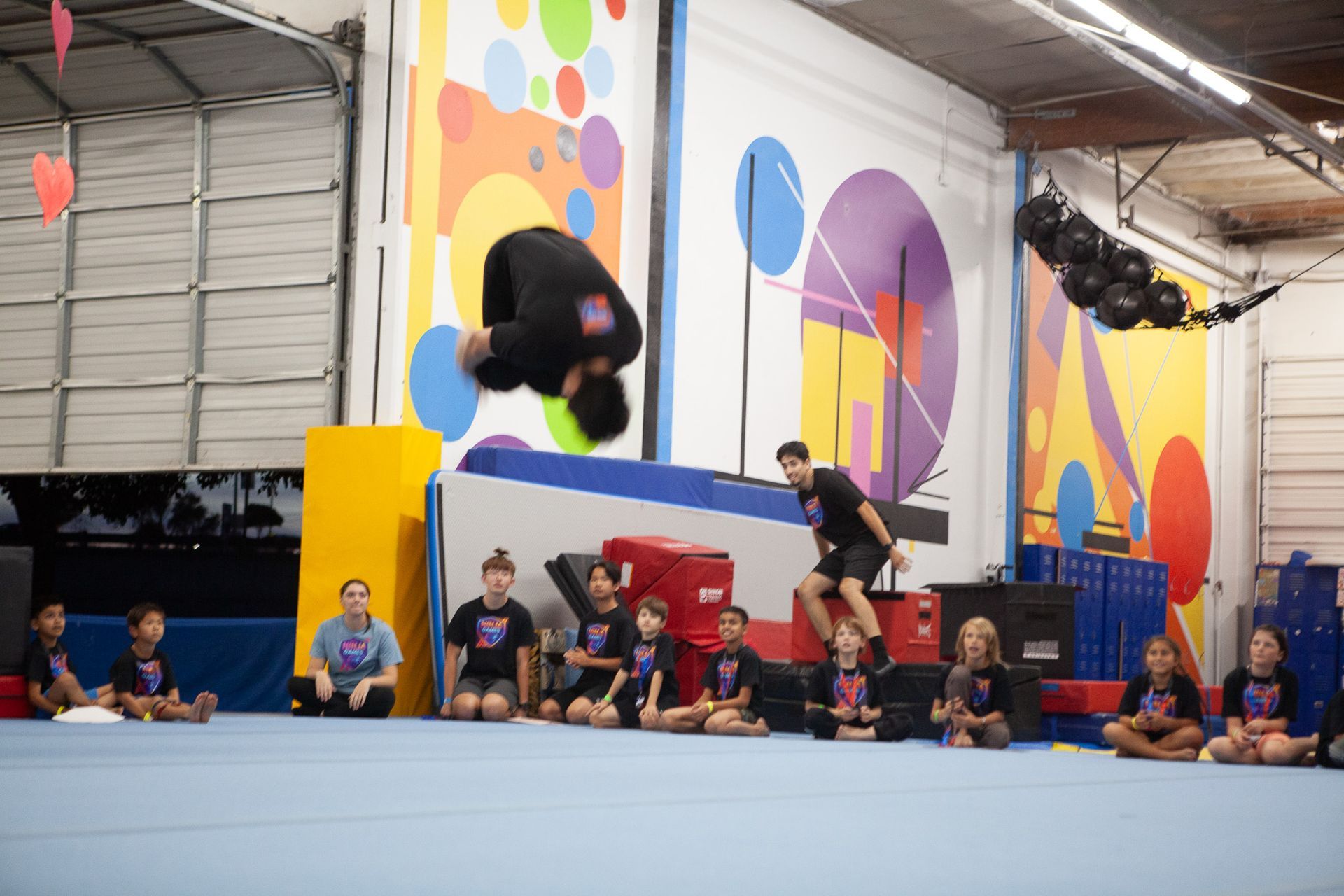 A person performs a backflip over a ledge in a gym; children watch.
