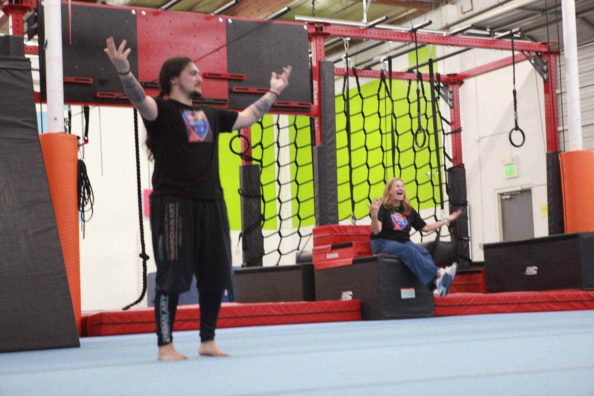 A man with tattoos stands with arms raised near a woman seated on a block, in a gym with obstacles.