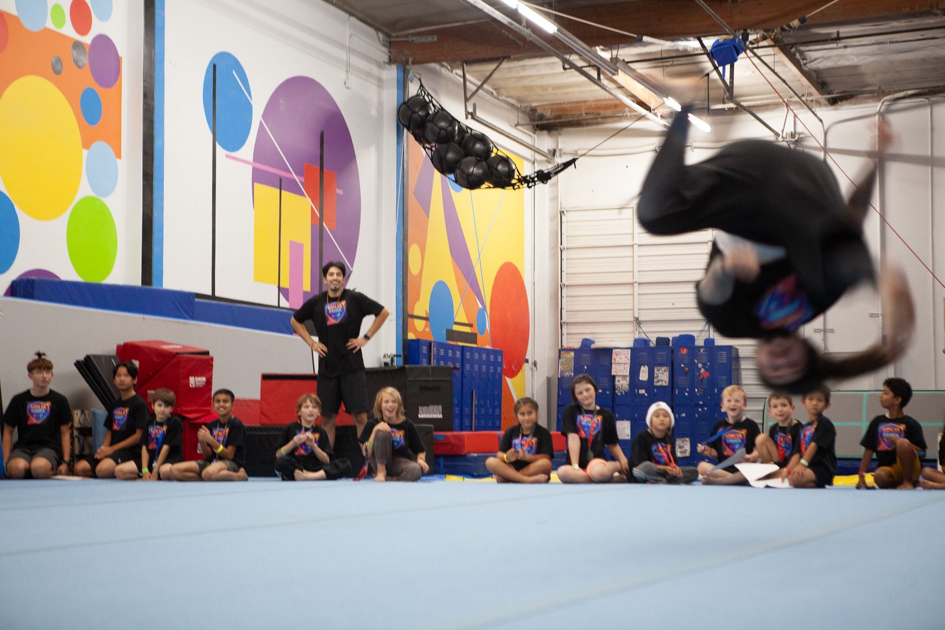 Person doing a backflip in a gym; a crowd of children watch. Colorful wall art behind.