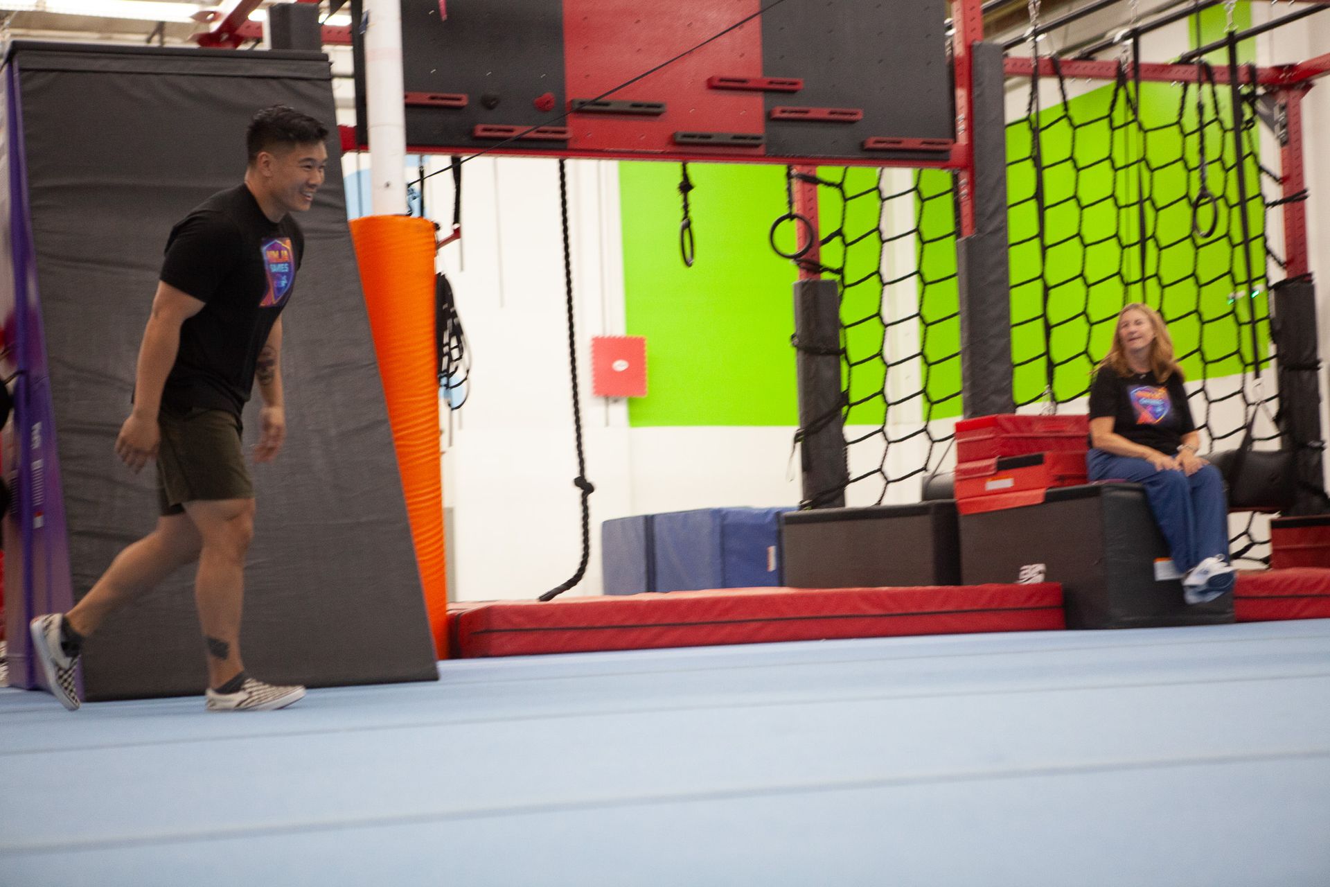 Man in black shirt and shorts preparing to run toward obstacle course, woman sitting nearby.