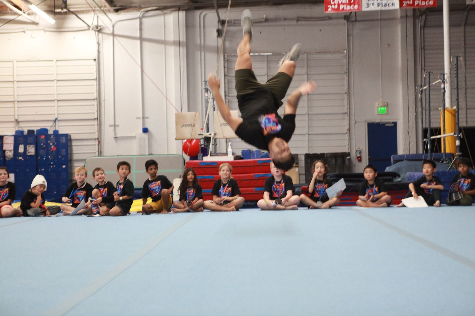 Man doing a backflip in a gym with children watching. Gym has blue mats and equipment.