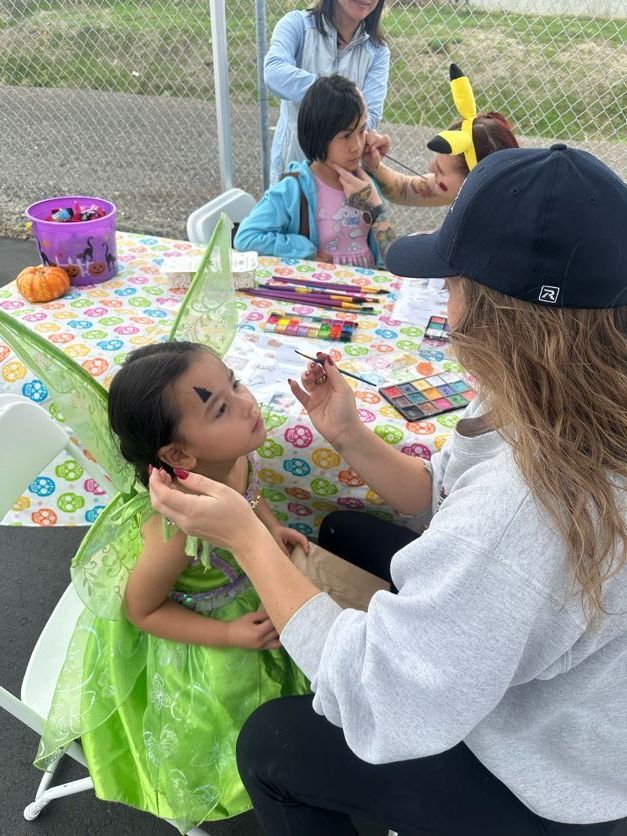 Girl in fairy costume having face paint applied at an outdoor event; other children watch.