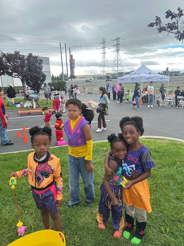 Children in colorful costumes at an outdoor event. Others watch nearby.