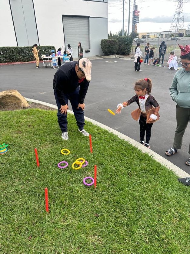 Child in costume throws ring at ring toss game. Adult watches. Outdoors.