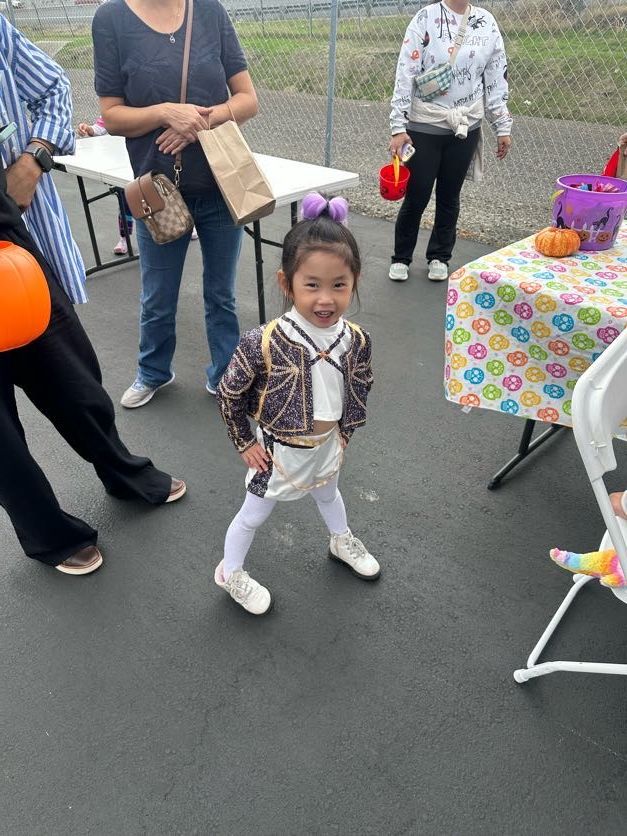 Young child in costume smiles, posing near a table with Halloween decorations; adults nearby.