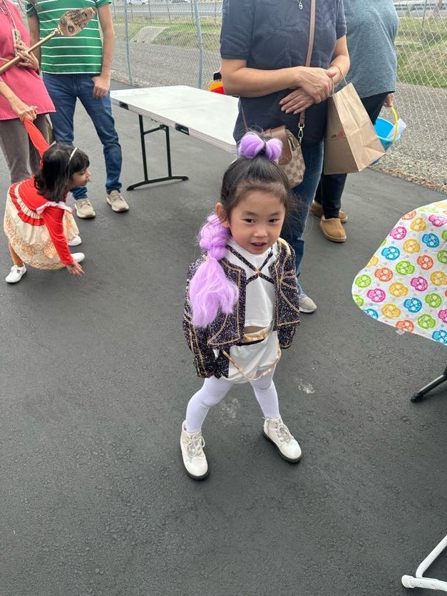 Girl in a costume, standing outdoors, looking at the camera. Others gather around a table.
