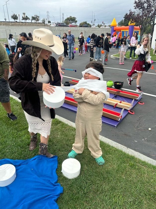 Woman in cowboy hat guides a blindfolded child at an outdoor event. They stand near craft supplies and colorful decorations.