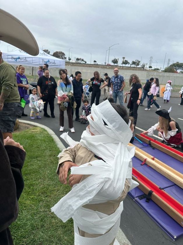 Child wrapped in toilet paper as a mummy at an outdoor Halloween event, others in costume in background.