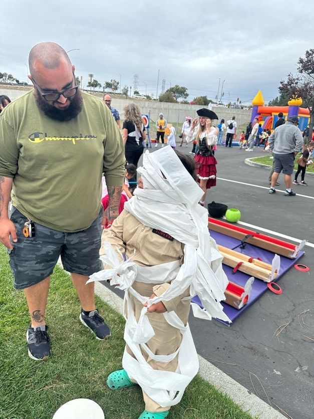 Man watches a child wrapped in toilet paper as a mummy costume at an outdoor event.