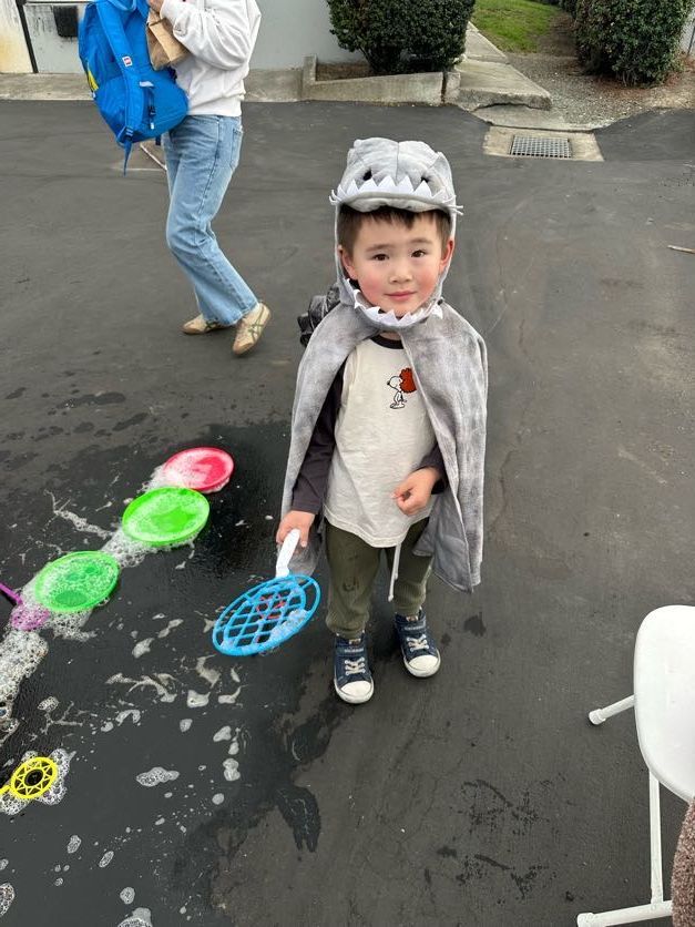 Young child in a dinosaur costume holds a bubble wand.  Bubbles and another person are nearby. Outdoors.