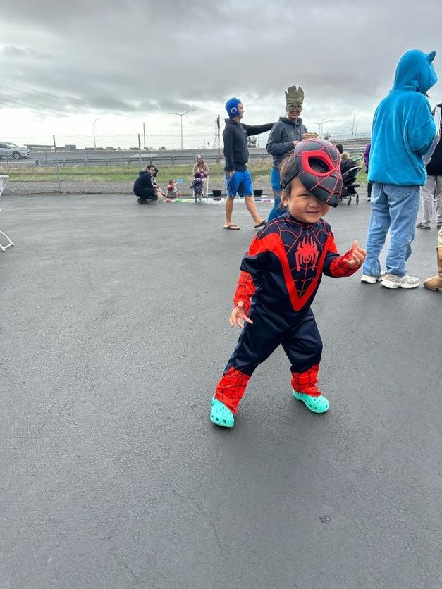 Toddler in Spider-Man costume, red and black, stands outside smiling. Gray overcast sky, people in background.