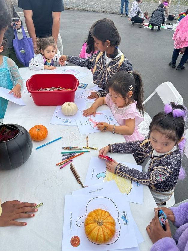 Children coloring pumpkin outlines at an outdoor table; pumpkins, crayons, and a red bin are present.
