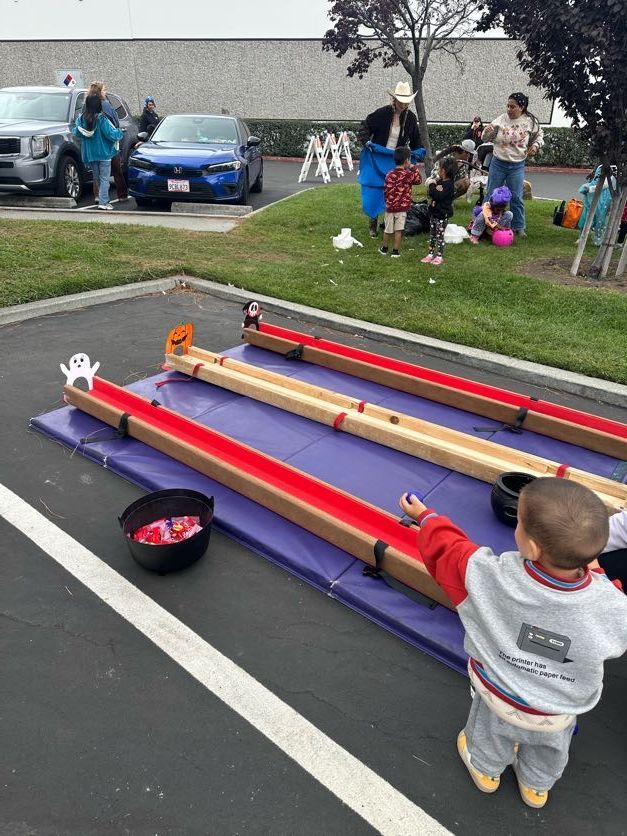 Child at a Halloween game, tossing objects down a wooden track. Spectators and cars in the background.
