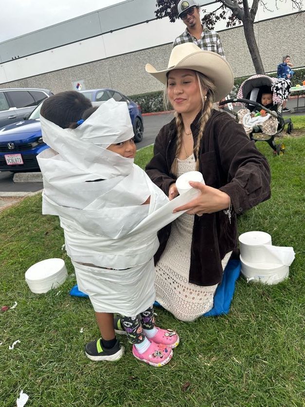 Person wrapping a child in toilet paper outdoors. Another person watches.