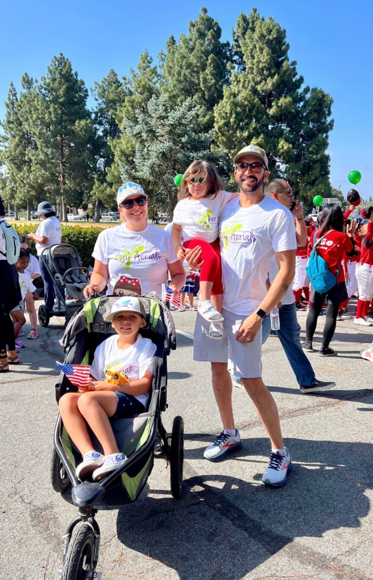 Family poses outdoors. A woman pushes a stroller. Other people visible, sunny.