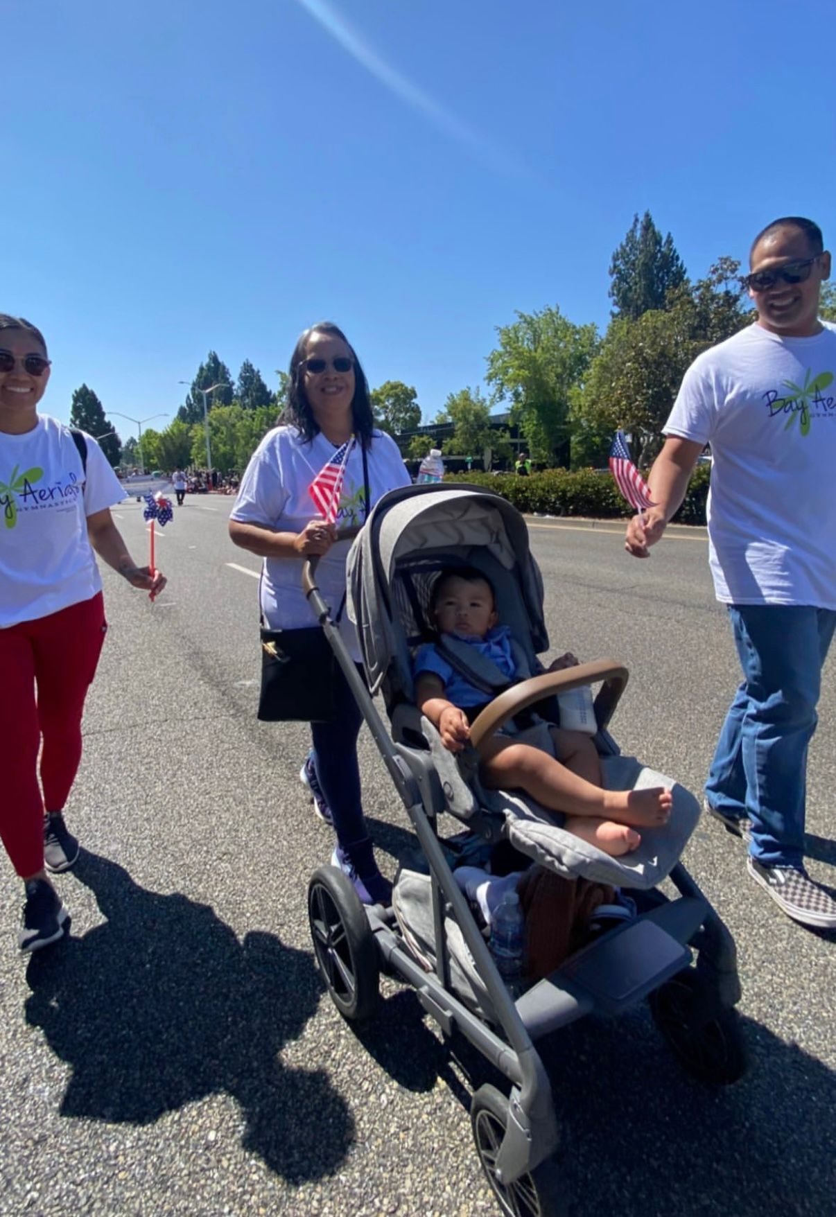 People walking in street parade, one pushing stroller with child. Holding American flags, sunny day.