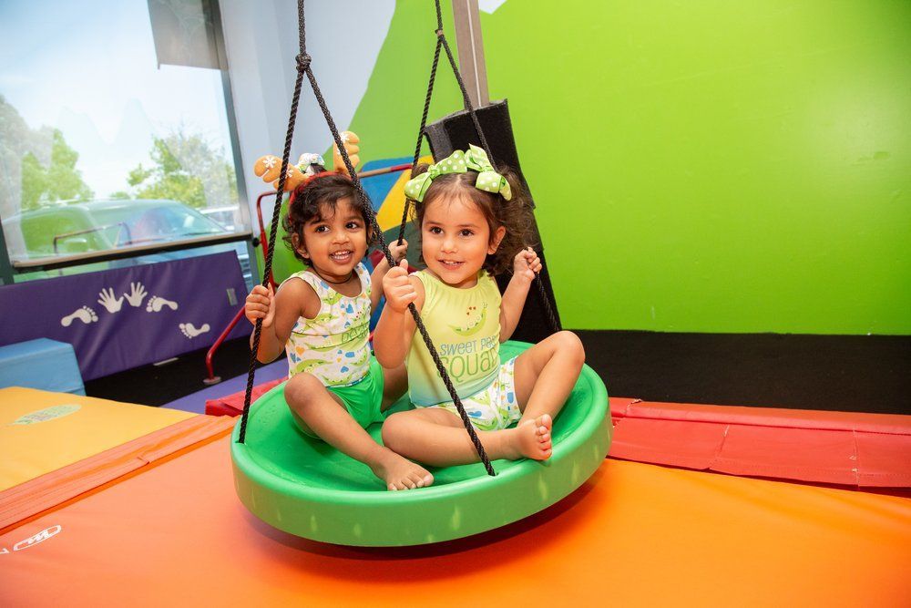 Two children on a green swing, smiling, in a brightly colored indoor play area.
