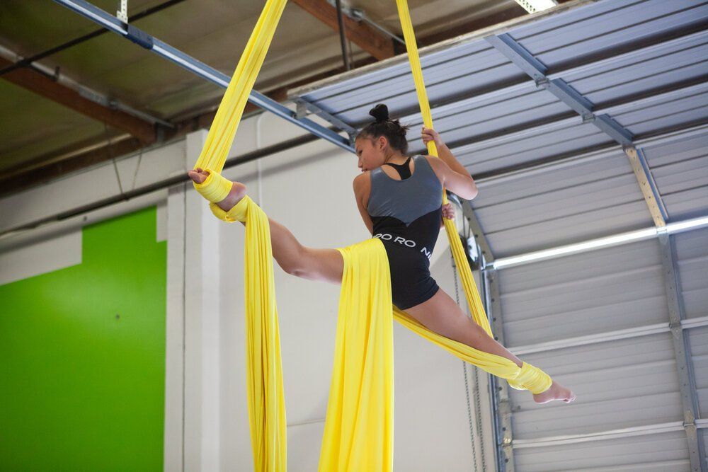 Woman in aerial silks, legs split, in a gym. Yellow fabric, black leotard, green and metal background.
