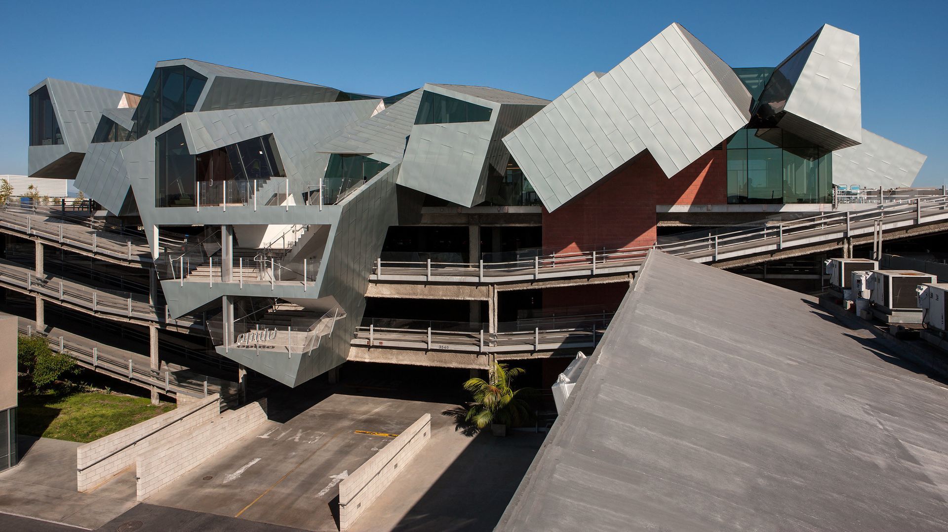 Modern angular building with metallic faceted roofs and wooden walkways under a blue sky