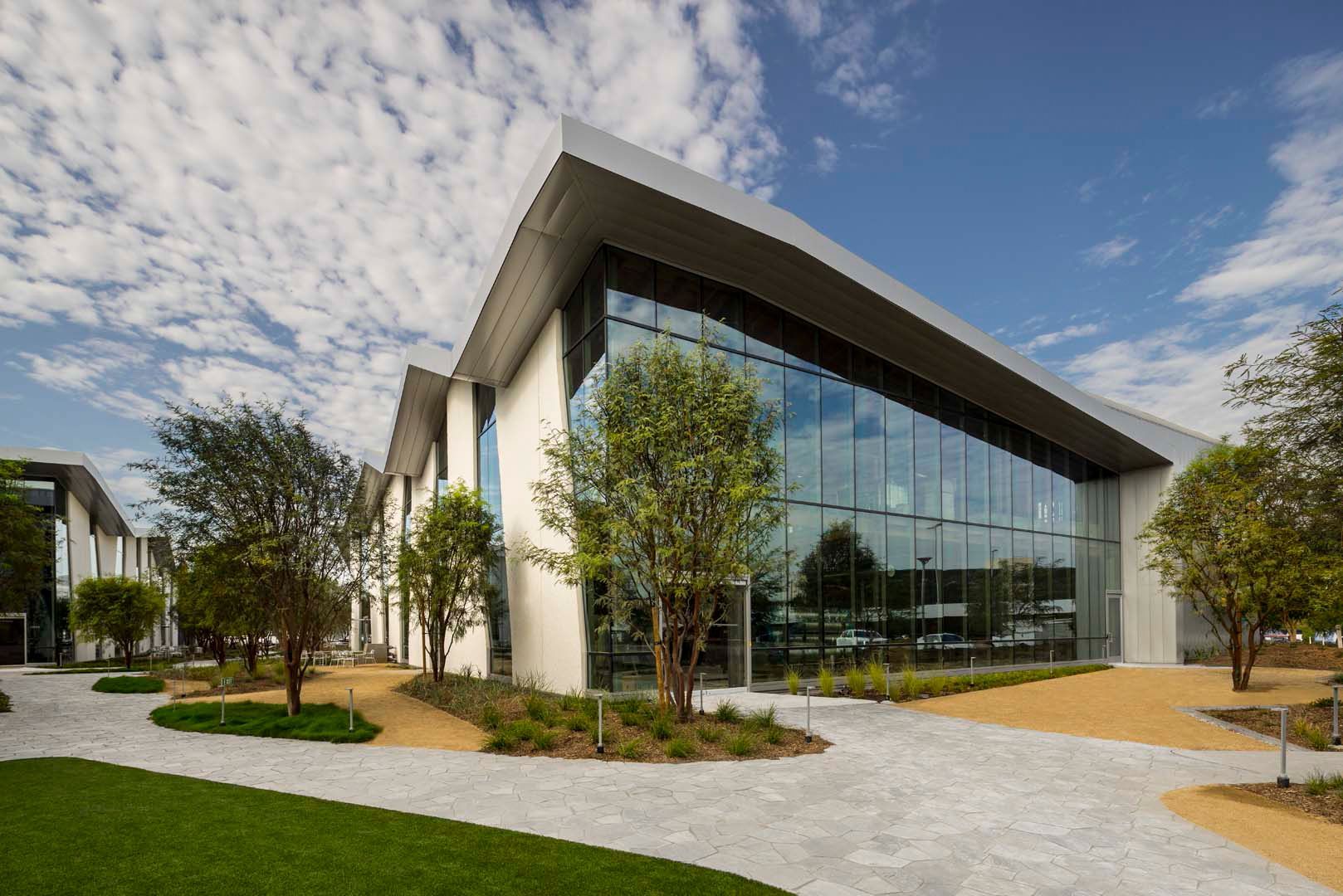 Modern glass office building with landscaped grounds and a curved walkway under a partly cloudy sky