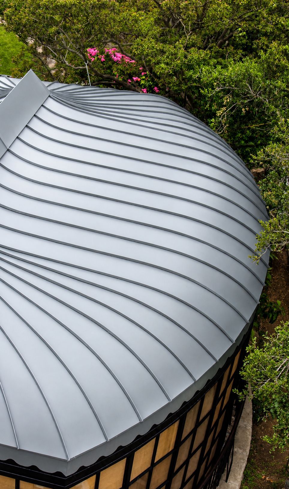 Curved gray metal roof beside lush green foliage and pink flowers