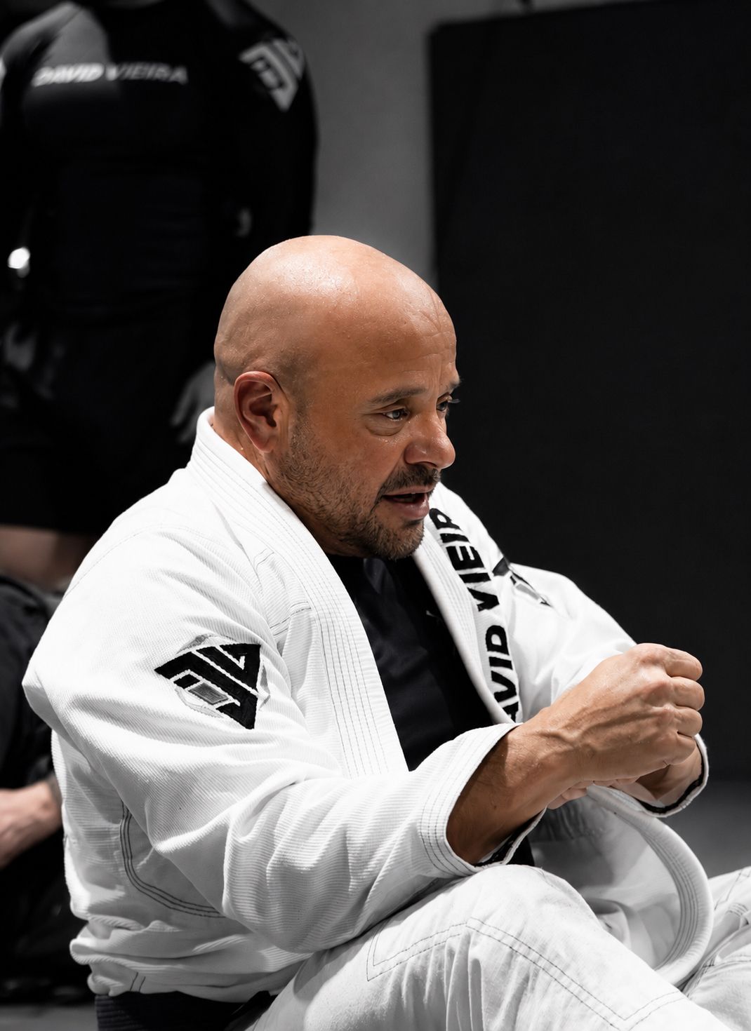Bald man in a white gi kneels in a martial arts gym, gripping his wrist in a focused pose