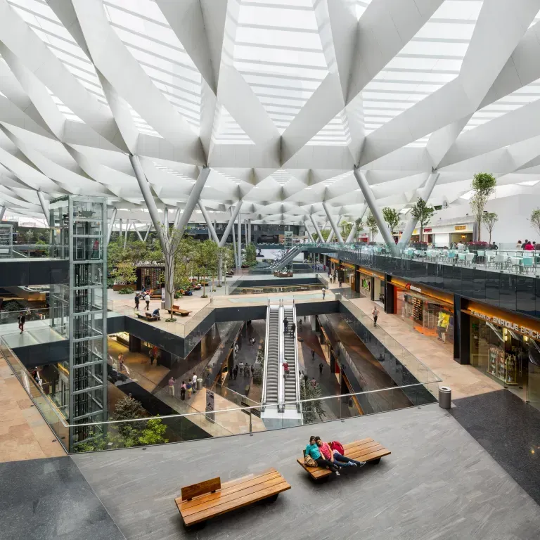 Modern indoor mall atrium with escalators, glass elevators, benches, and white geometric ceiling