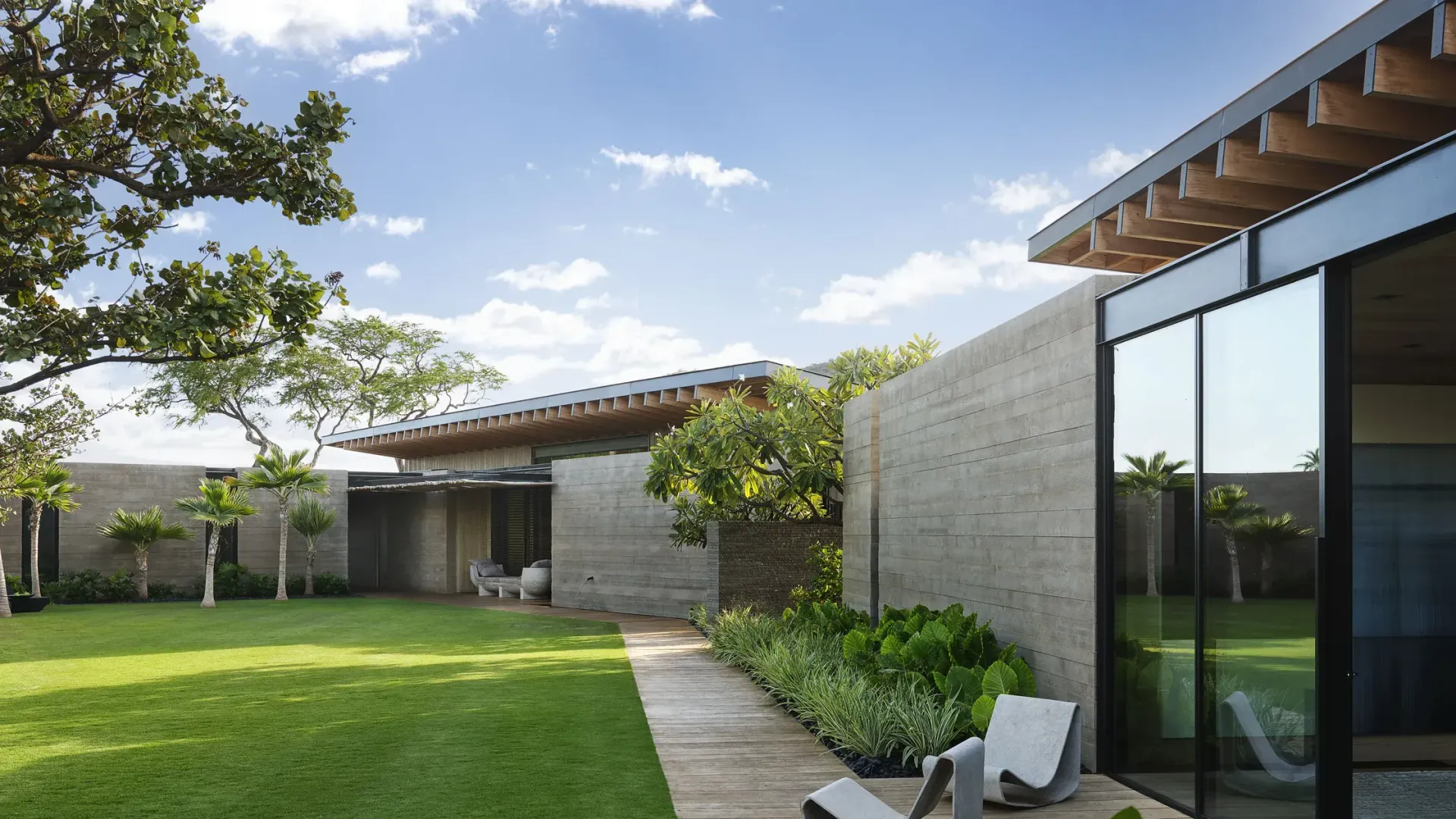 Modern courtyard with green lawn, stone path, trees, and a glass-walled building under a blue sky.