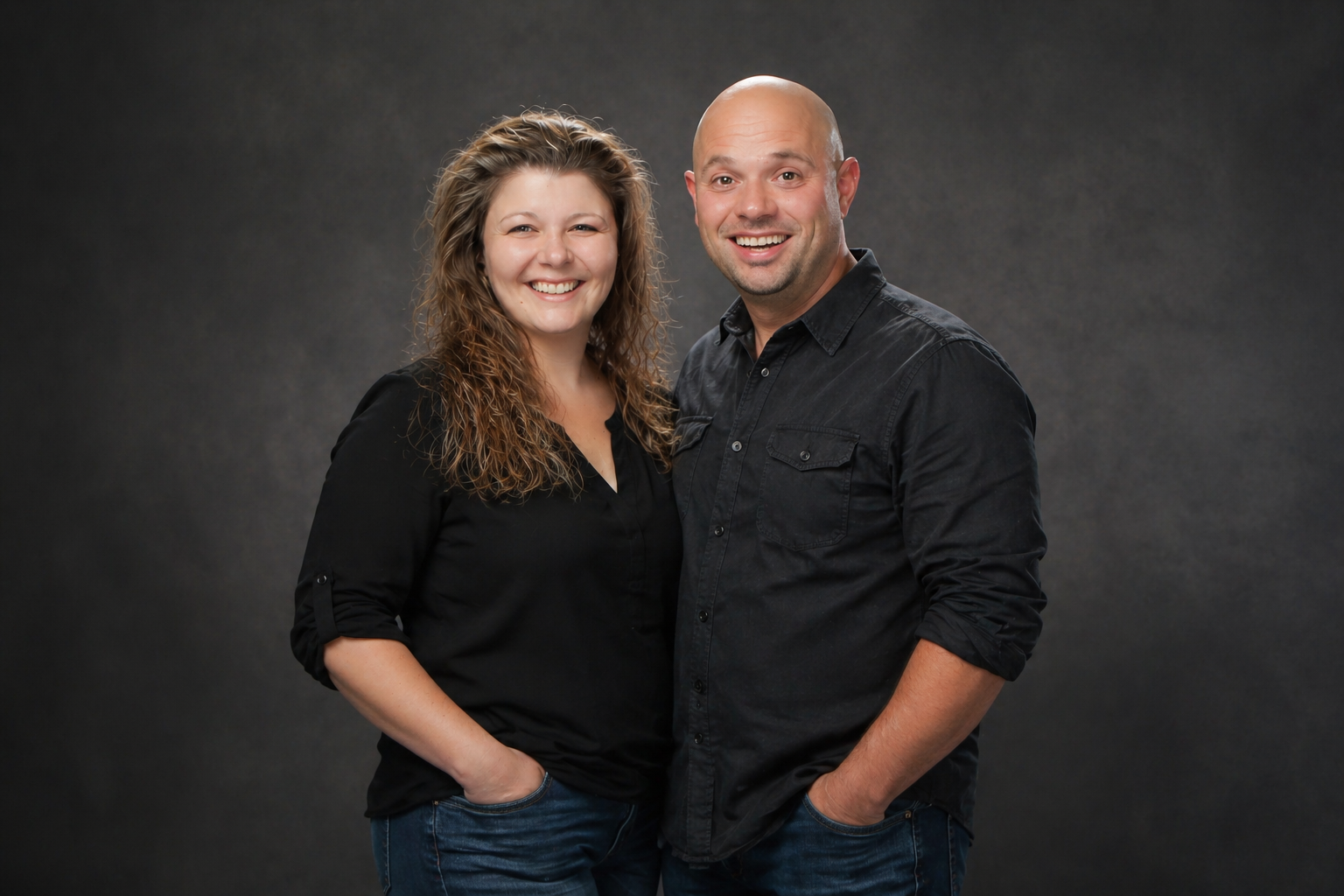 Two people standing side by side, smiling, against a dark studio backdrop.