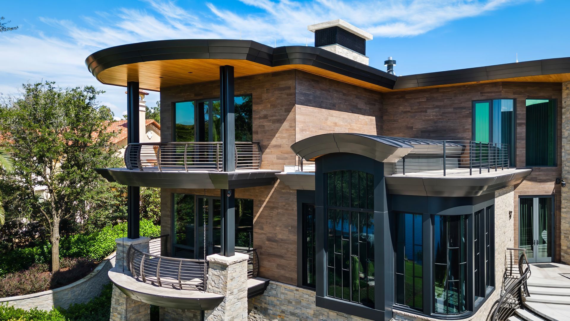Modern two-story house with curved balconies, large glass windows, and brick and wood exterior, surrounded by trees