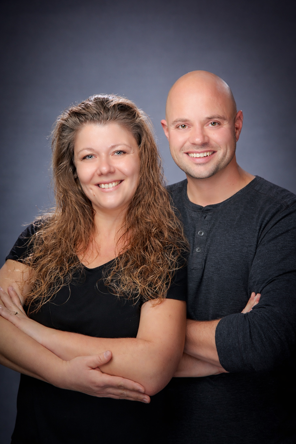 Smiling couple posing together against a gray studio backdrop, arms wrapped around each other.