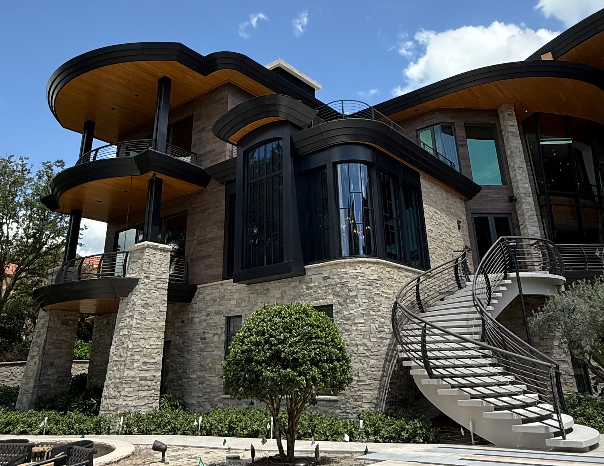 Modern stone house with curved balconies, large windows, and a spiral staircase under a blue sky