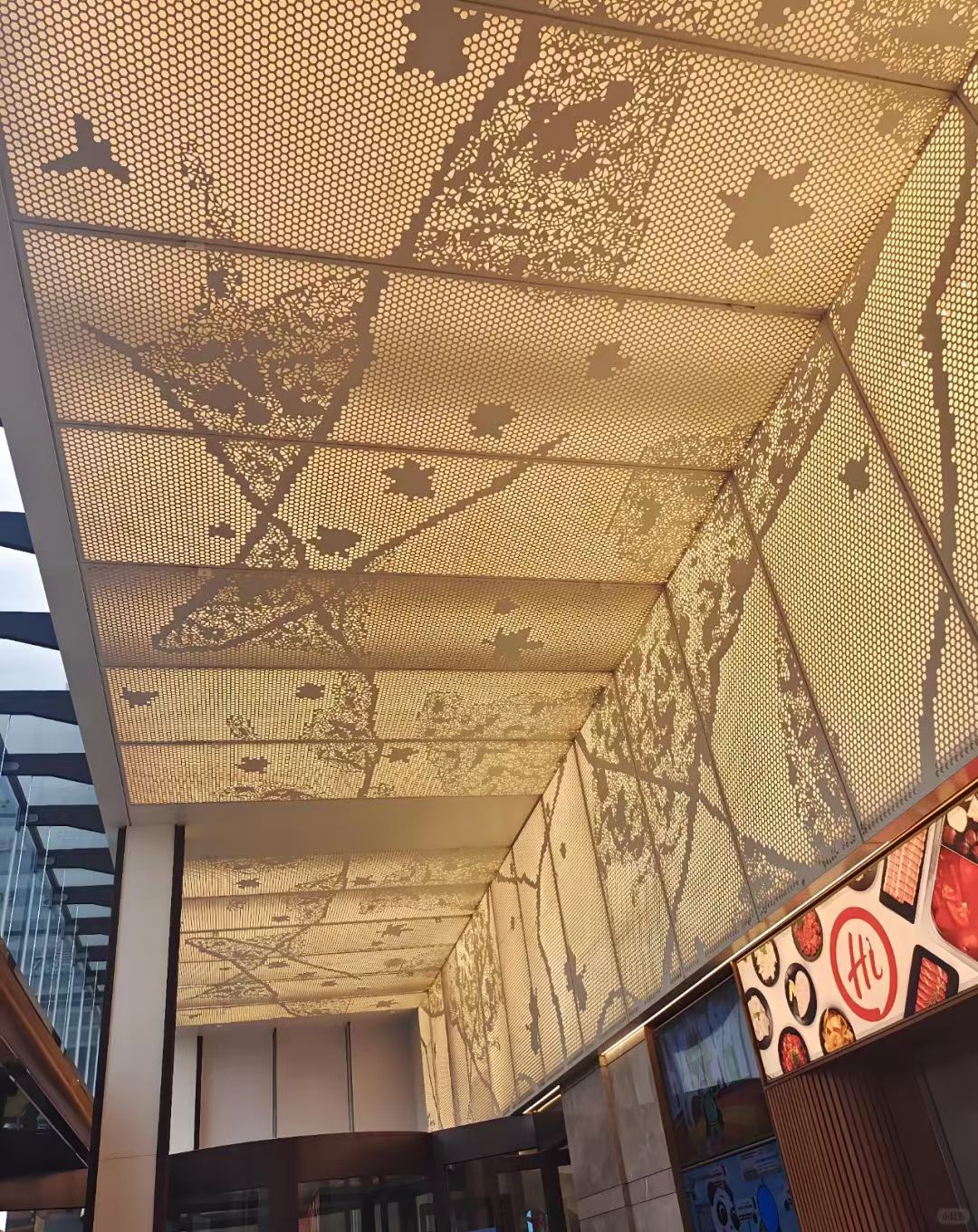 Decorative perforated metal canopy casting leaf-like shadows over a covered walkway with storefront signs below
