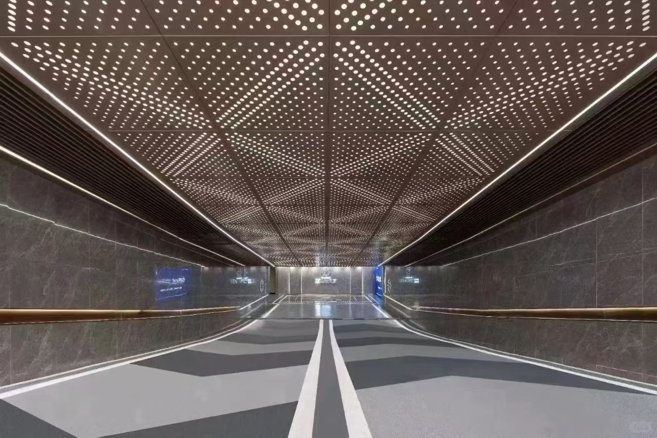 Modern pedestrian tunnel with perforated metal ceiling and walls, lit by rows of small lights.
