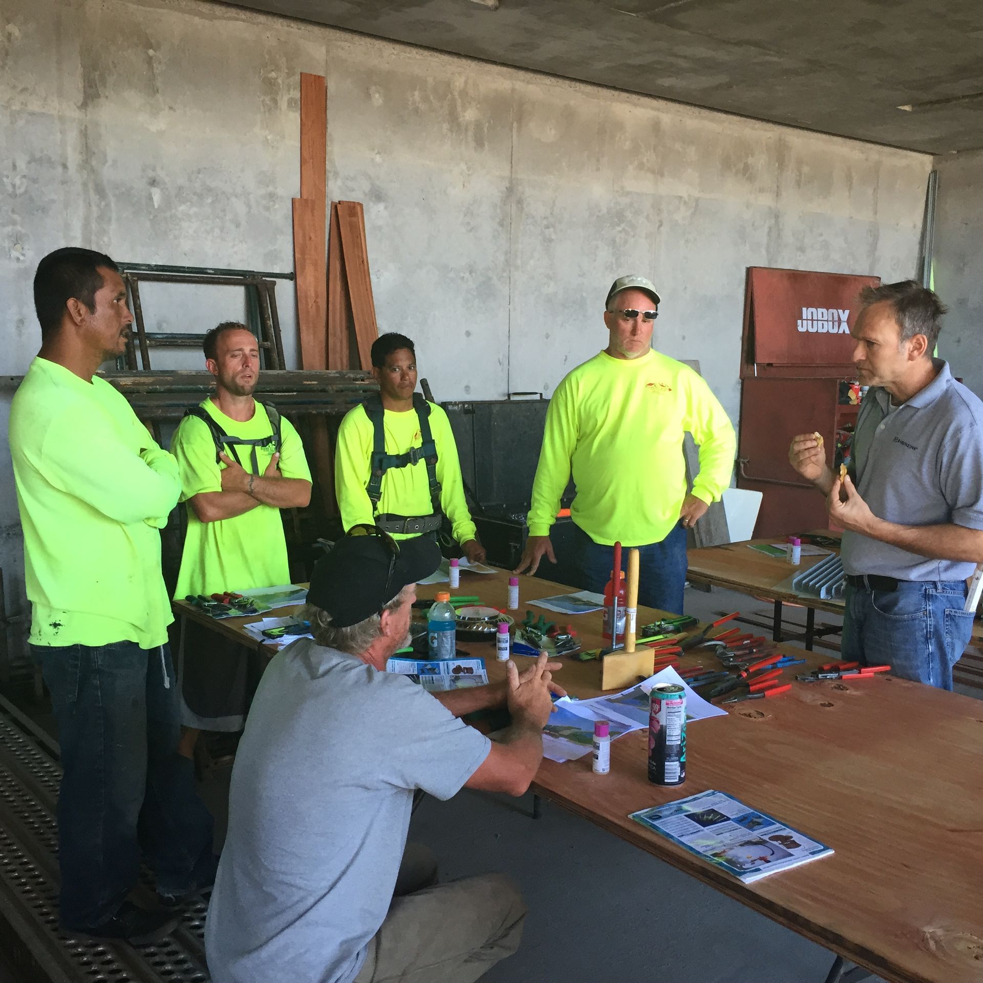 Workers in neon safety shirts gather around a table in a concrete room for a briefing.