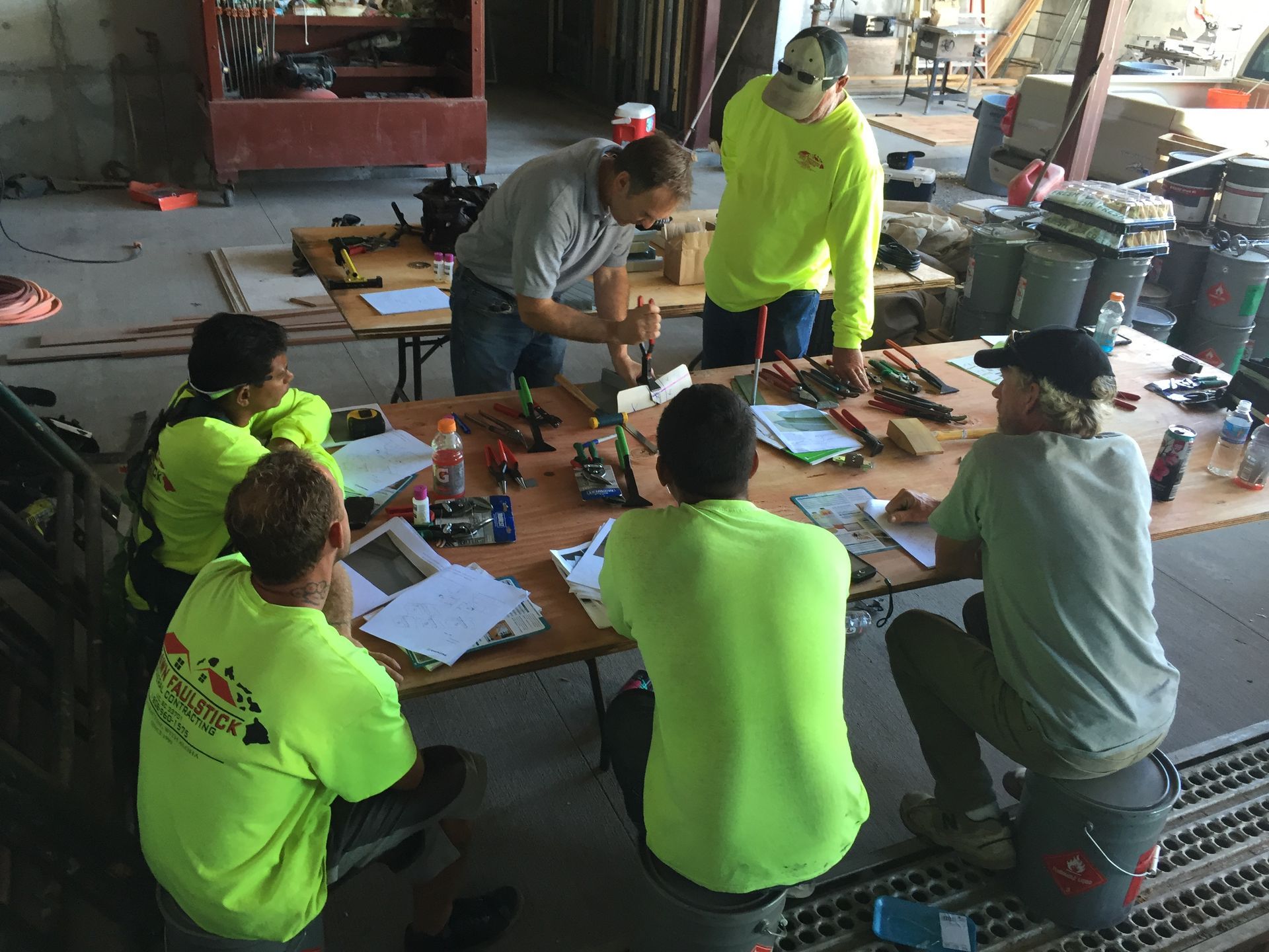 People in bright neon shirts gathered around a table in a workshop, discussing papers and tools.