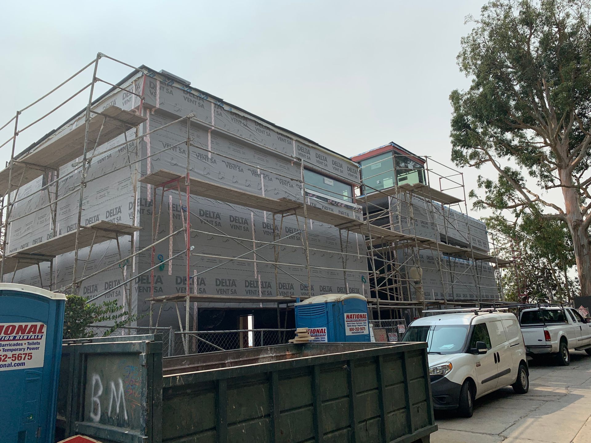 Scaffolded building under construction beside parked vans and a large dumpster on a street