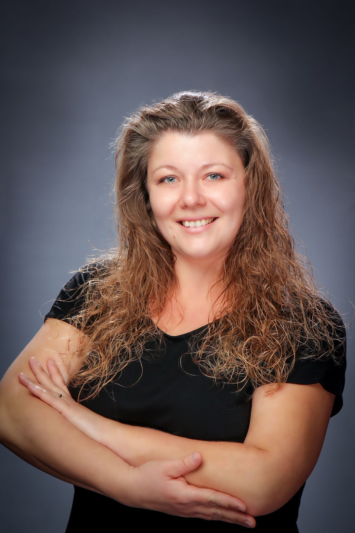 Smiling person with long wavy hair and black shirt, arms crossed against a gray studio backdrop