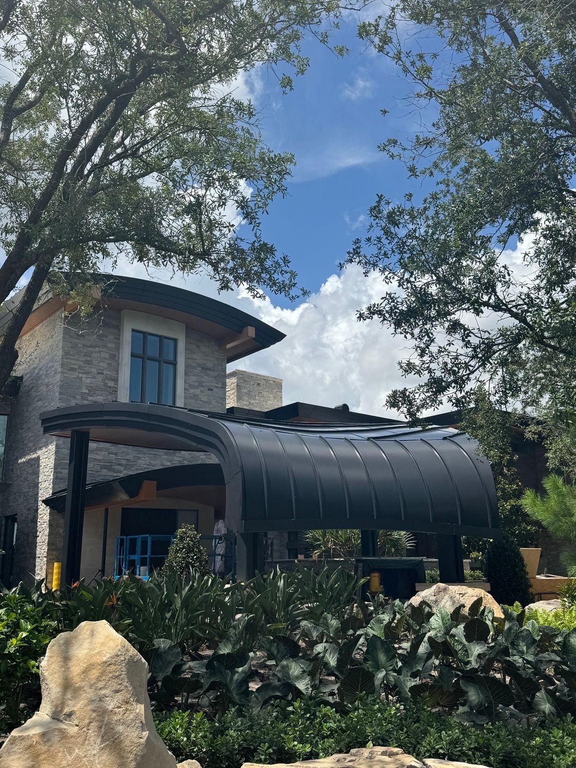 Stone house with dark awning, surrounded by trees and greenery under a blue sky