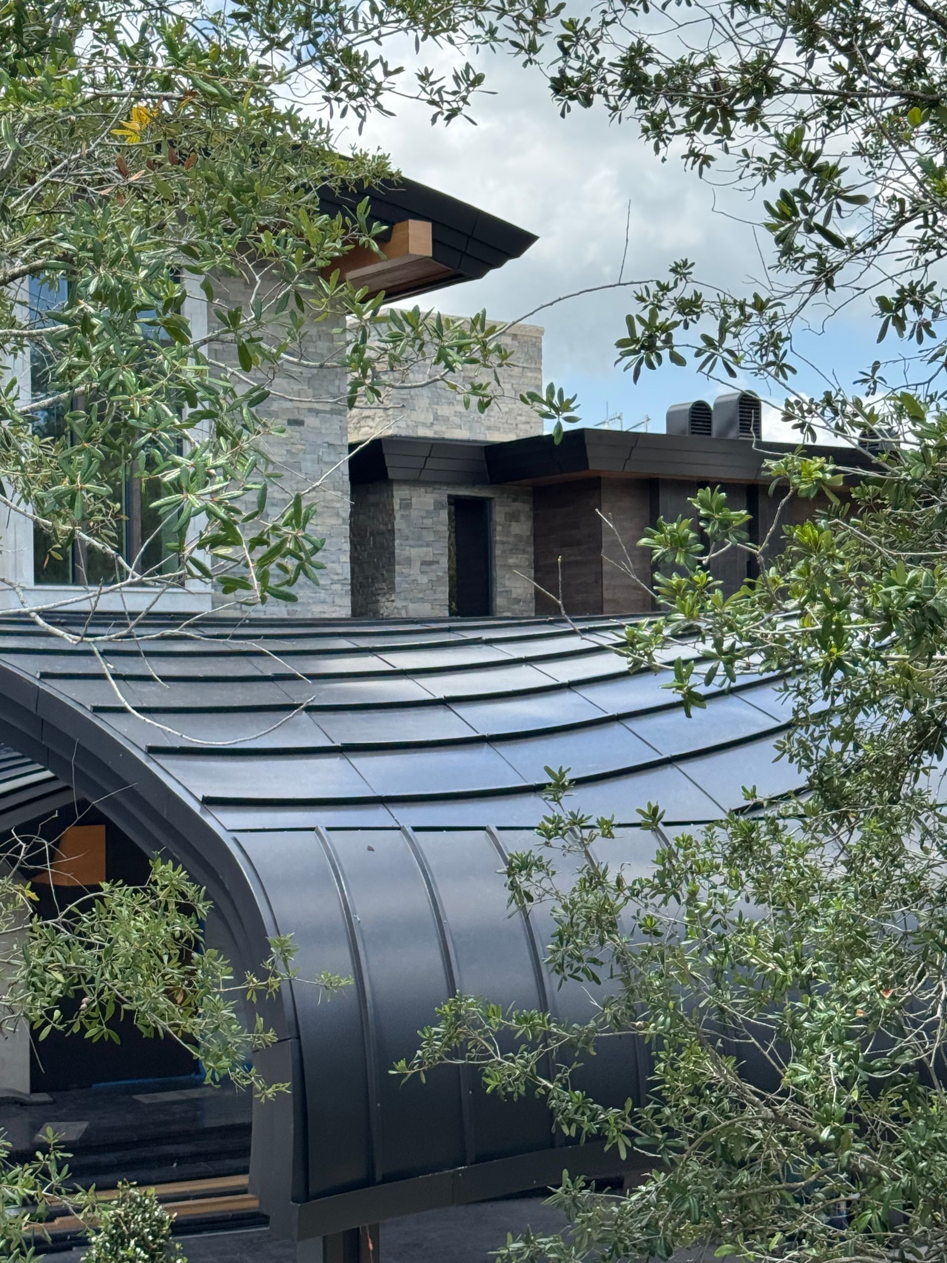 Modern house with dark metal roof and stone chimney, framed by leafy trees under a cloudy sky