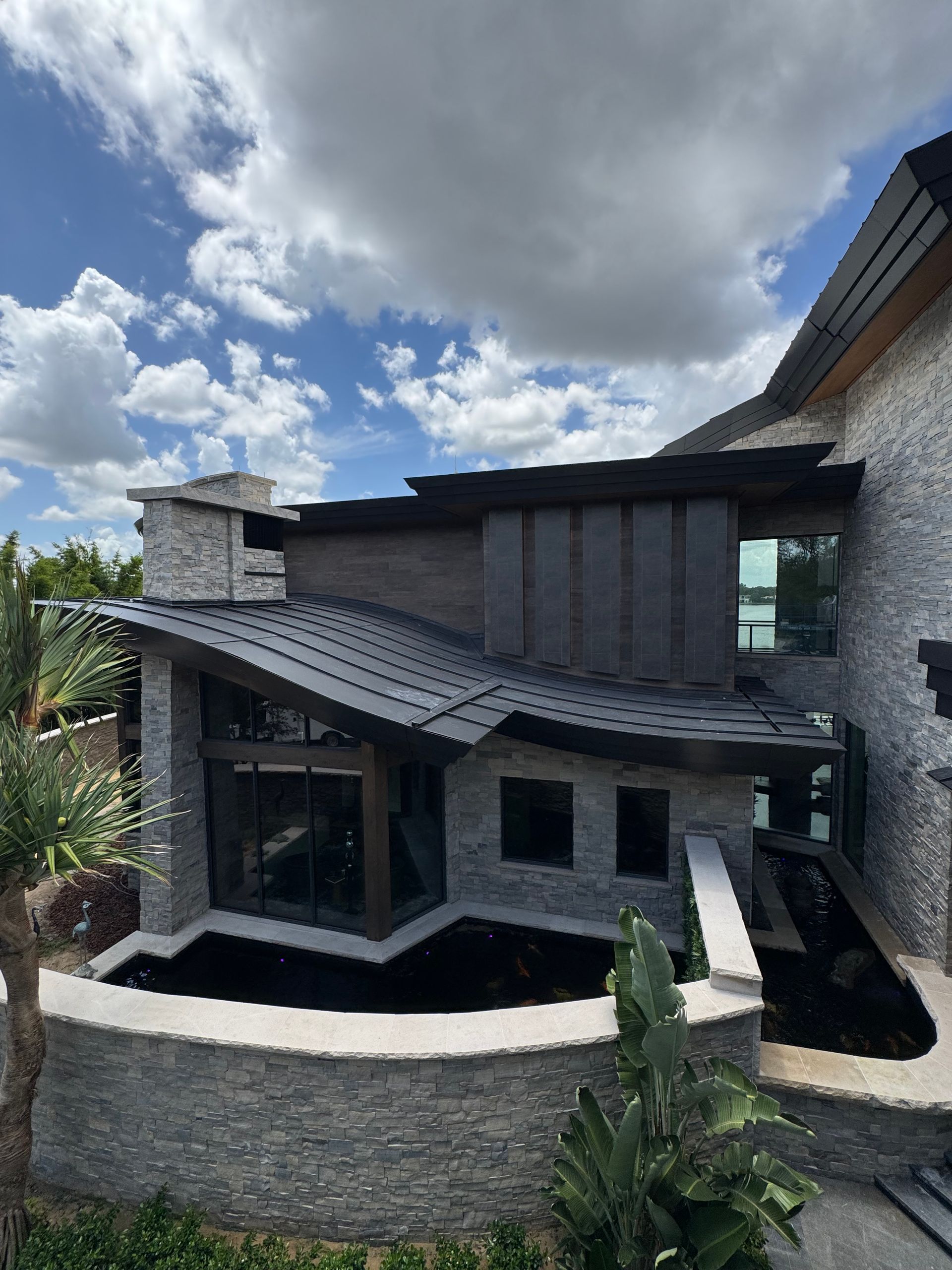 Modern stone house with dark roof, large windows, and landscaped yard under a cloudy sky