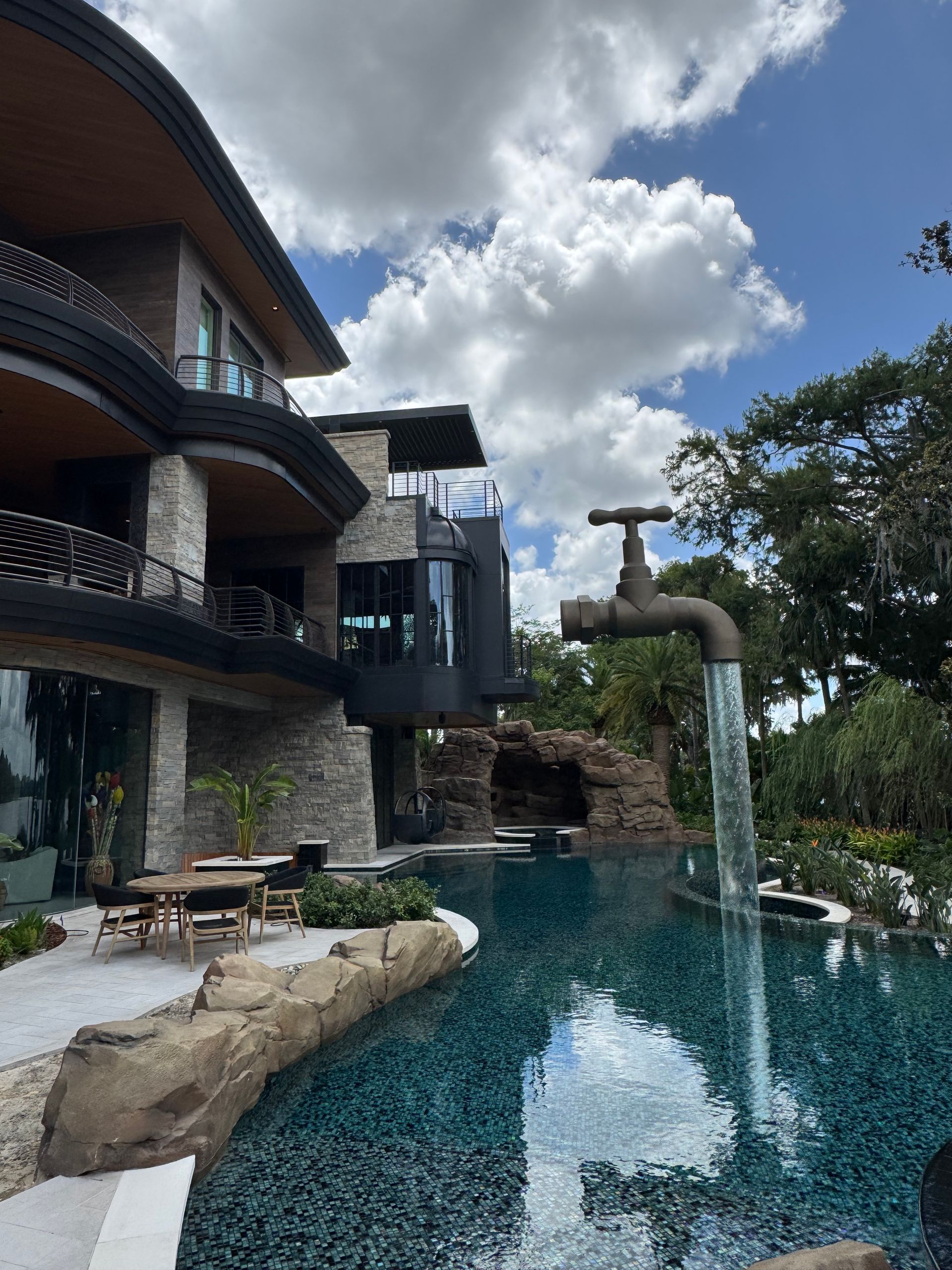 Resort pool with a waterfall, stone building balconies, and blue sky with clouds