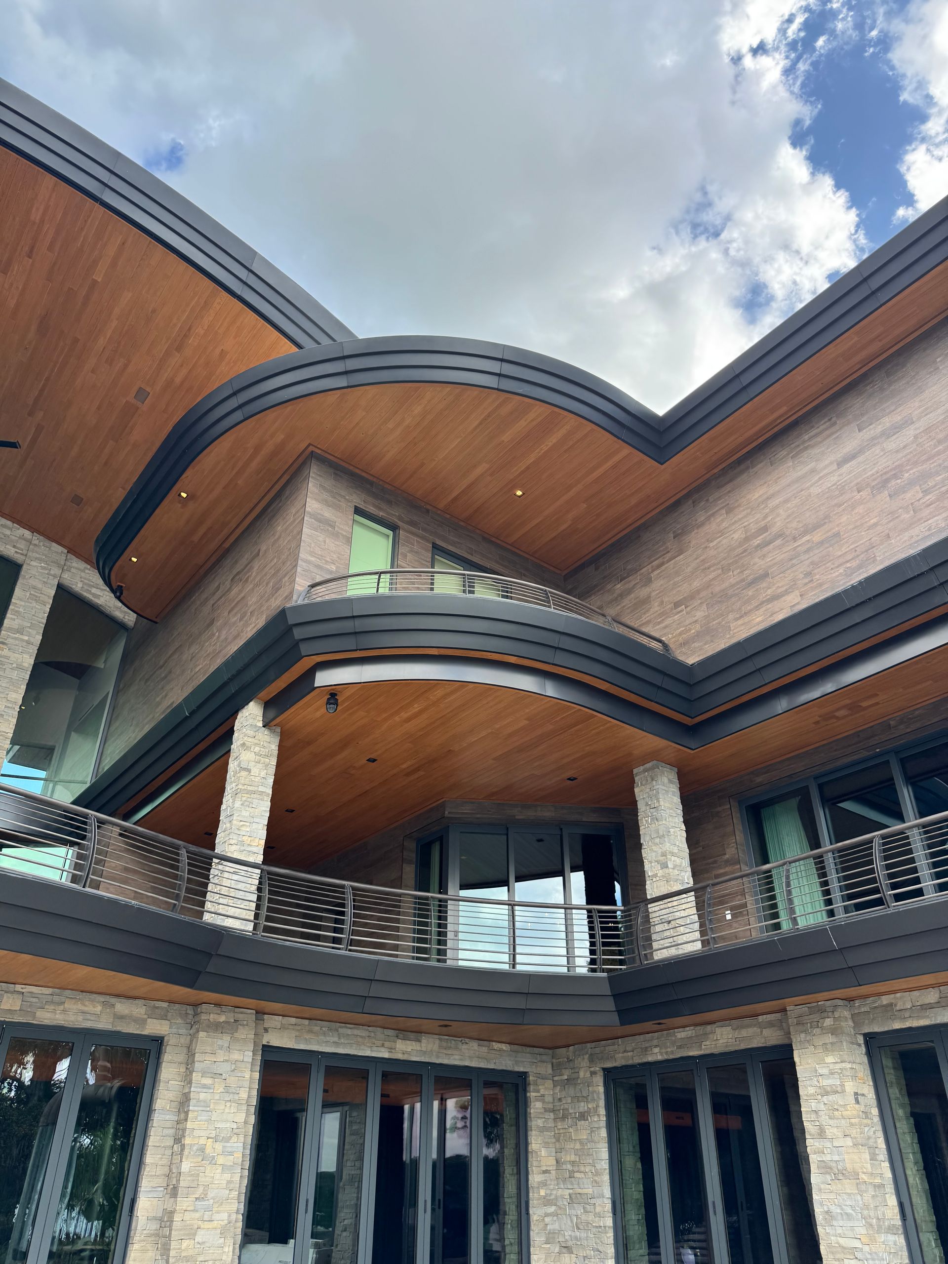 Modern multi-story house with curved wood-paneled roof and brick balconies under a cloudy sky