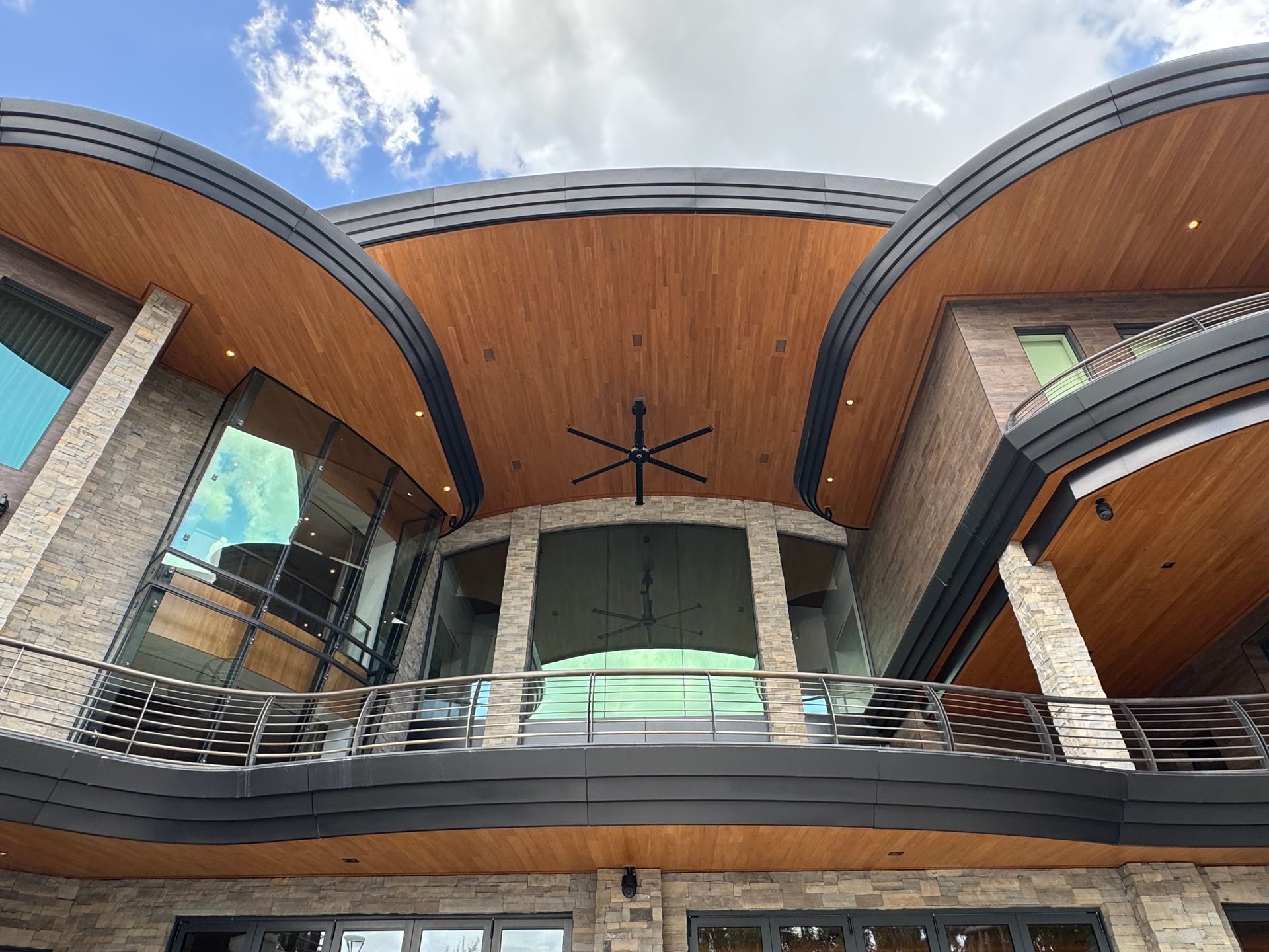 Modern building exterior with curved wood ceiling, brick columns, glass railings, and blue sky.